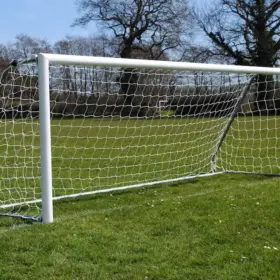 A white soccer goal with netting stands on a grassy field under a clear blue sky. Leafless trees surround the field, creating a calm, open atmosphere.