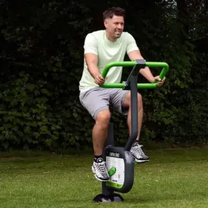 A man on an outdoor exercise bike in a park, wearing a light green shirt and gray shorts. The scene is set against lush green trees, conveying a calm, active vibe.