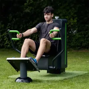A man exercises on a row machine outdoors. He wears a grey T-shirt and black shorts, with a focused expression. Green grass and bushes are in the background.